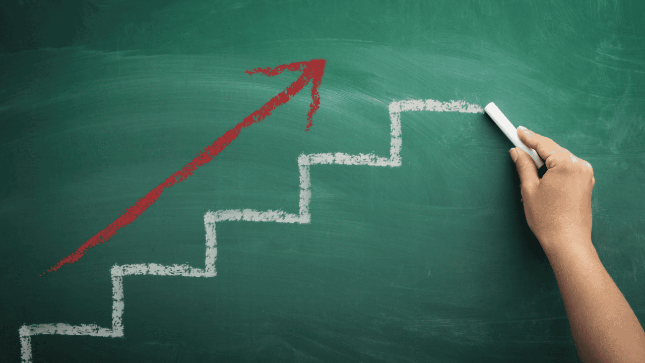 Hand drawing a white staircase with an upward-pointing red arrow on a green chalkboard, illustrating incremental progress and small steps toward success.