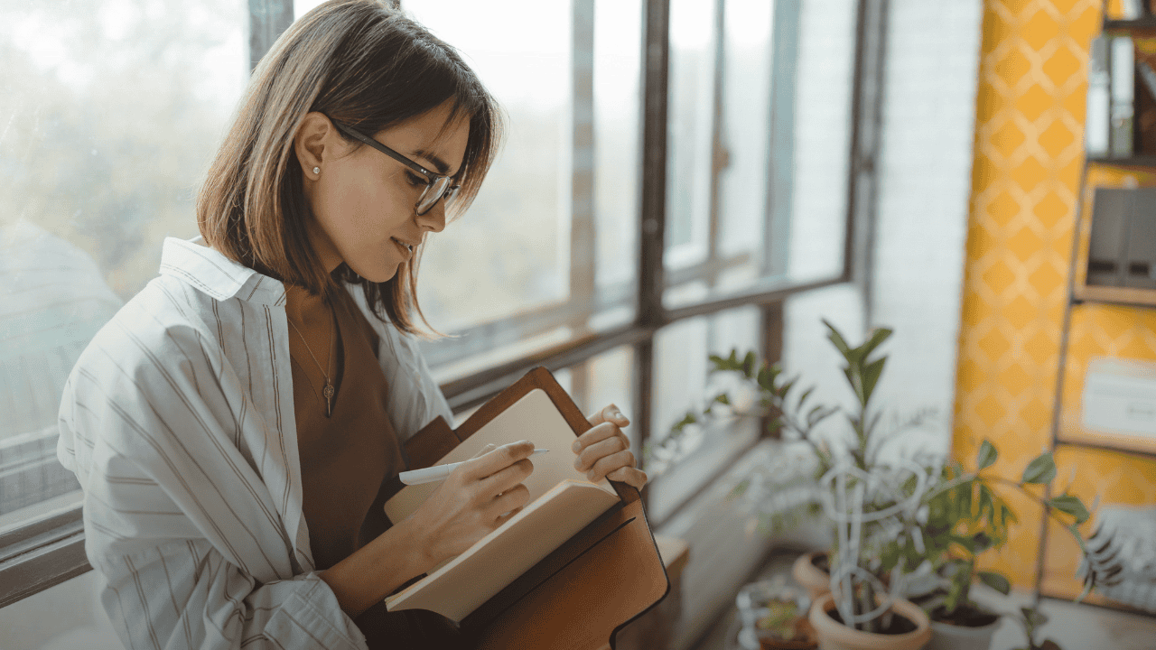 Woman standing by a sunlit window calmly writing in a leather notebook, depicting mindful reflection and stress-free goal setting for the new year.