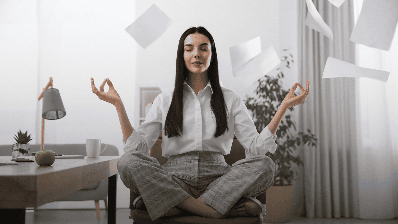 Self-Care in High-Stress Workplaces Businesswoman meditating in an office chair with papers flying around her, demonstrating how to maintain mindfulness and focus in a chaotic, high-pressure work environment.