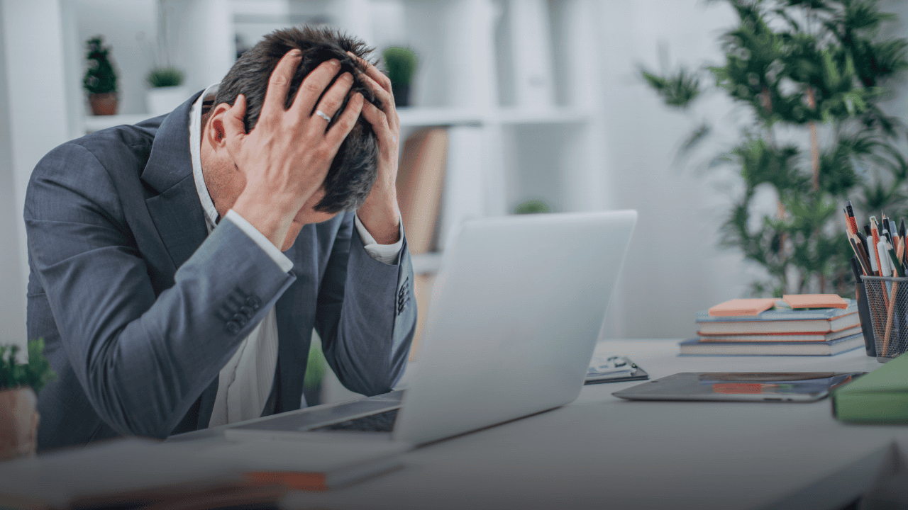 Stressed man holding his head in his hands at an office desk, depicting the isolation and hidden distress caused by the stigma around mental health at work.