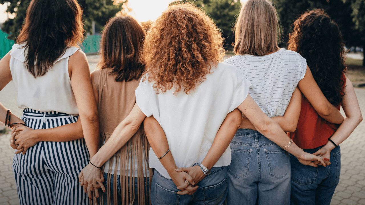 A diverse group of women standing in a row with arms linked and hands held, symbolizing the strong foundation and collective power required to build a shared future for International Women’s Day.