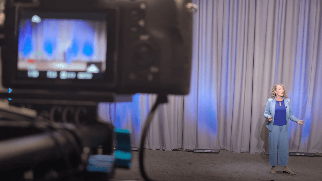 Tammy Ward speaking on a professional stage during a keynote event, captured through the lens of a video camera, discussing leadership and growth through change.