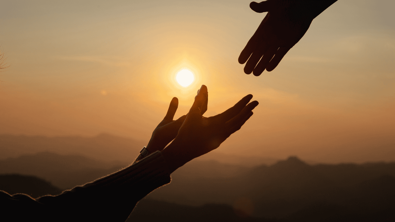 Silhouette of two hands reaching toward each other against a warm sunset over a mountain horizon, representing hope, reinvention, and the strength found in human connection.