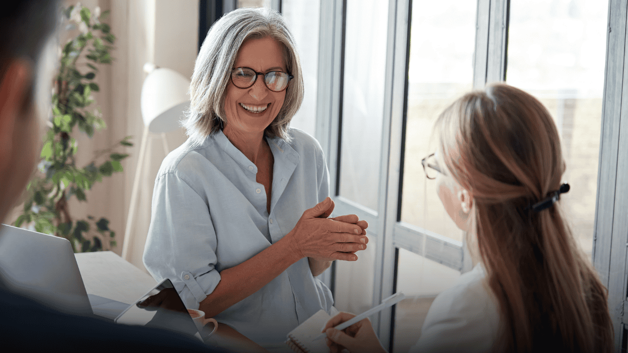 A smiling mature female leader in glasses actively engaging and speaking with a colleague during a workplace meeting, demonstrating authentic human leadership, warmth, and mentorship.