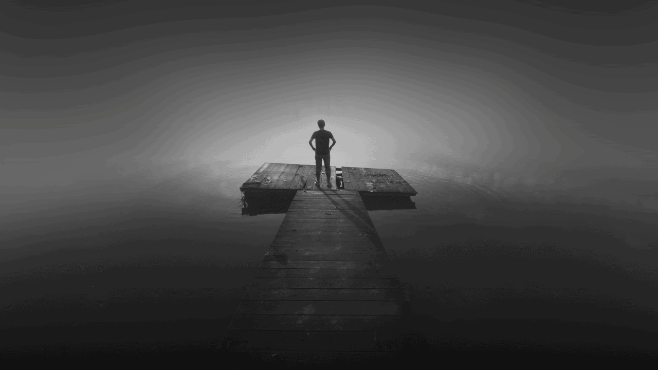 A silhouette of a person standing alone at the edge of a wooden dock extending into a foggy, misty water landscape, representing isolation, trauma, and the silent mental health struggles