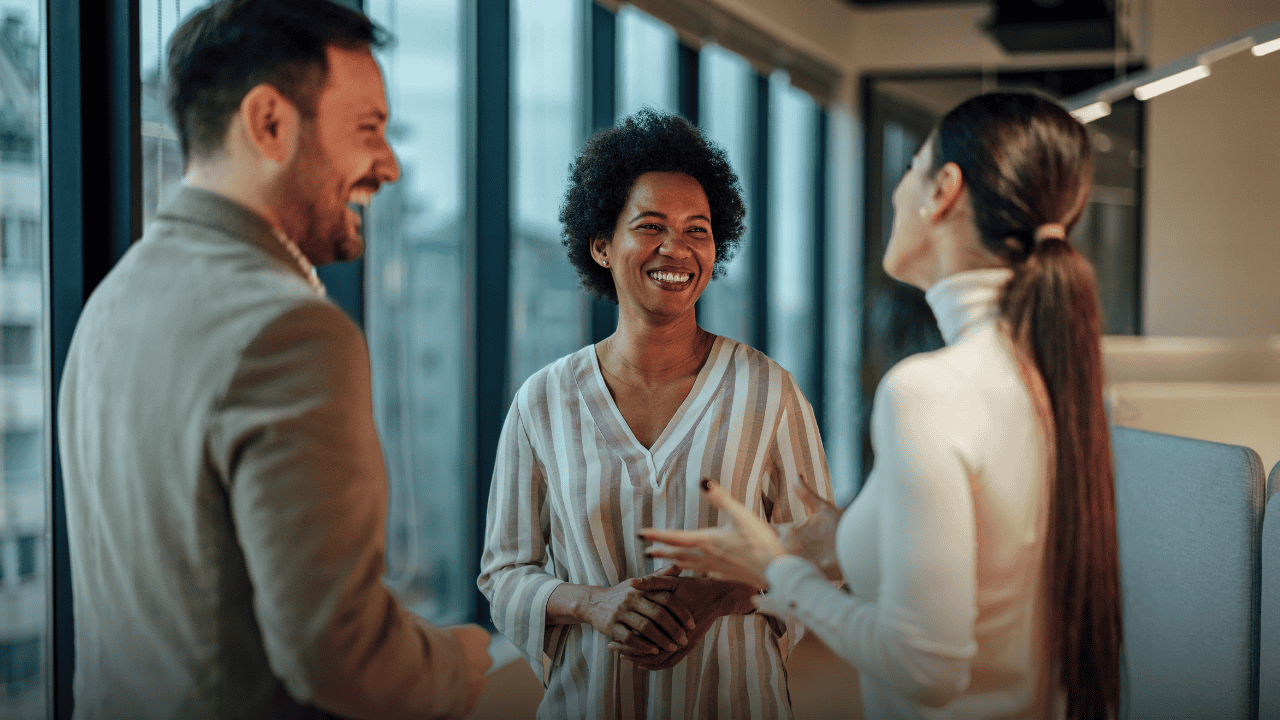 Three diverse corporate colleagues engaging in warm, open conversation in a bright office environment, representing how supportive leaders foster psychological safety and everyday workplace mental health connection.